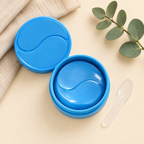 Open jar of blue collagen gel eye patches with spatula and eucalyptus on a cream background